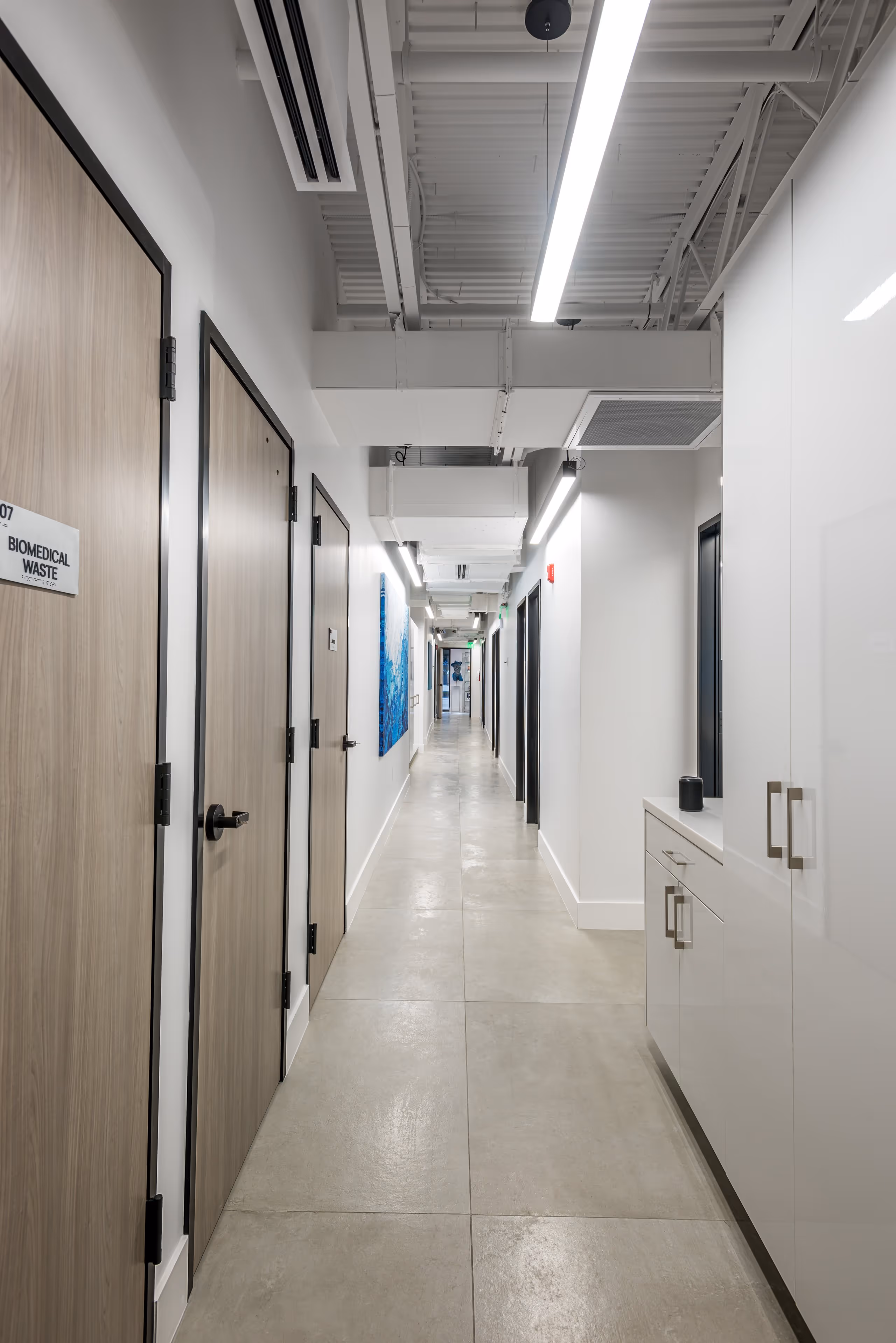Long, clean medical hallway with light wood doors on the left, white cabinets on the right, and bright overhead lighting.