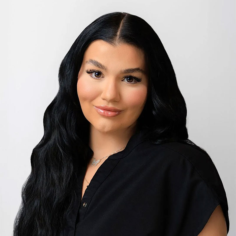 Woman with long black hair wearing a black top and a necklace, smiling against a white background.