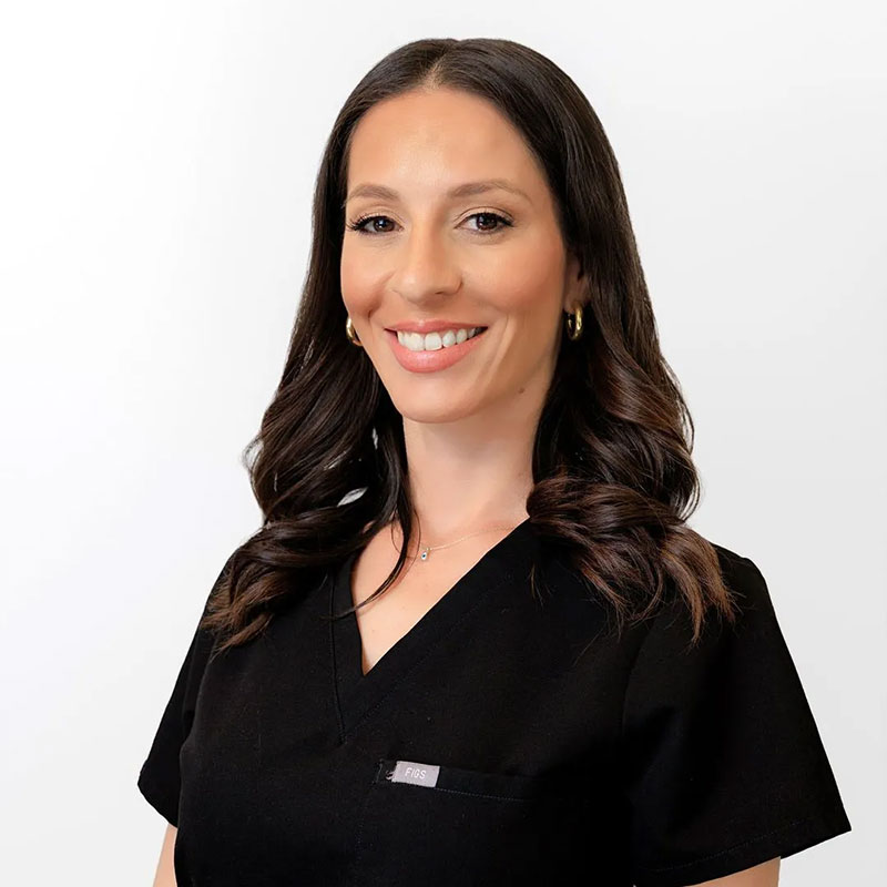 Smiling woman with long dark hair wearing black medical scrubs against a white background.