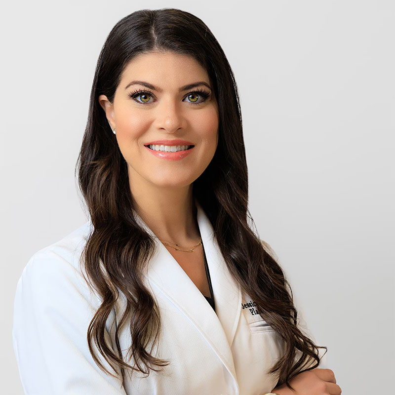 Smiling woman with long dark hair in a white medical coat against a light gray background.