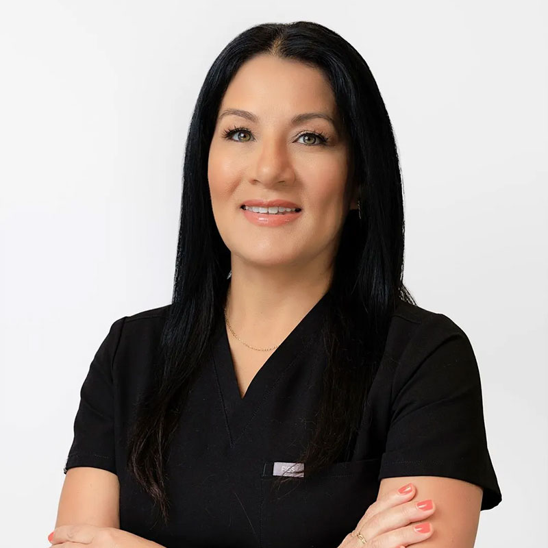 Smiling woman with long black hair wearing black medical scrubs with arms crossed on white background.