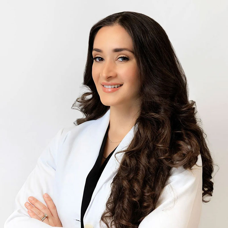 Woman with long curly dark hair wearing a white coat smiling with arms crossed in front of a white background.