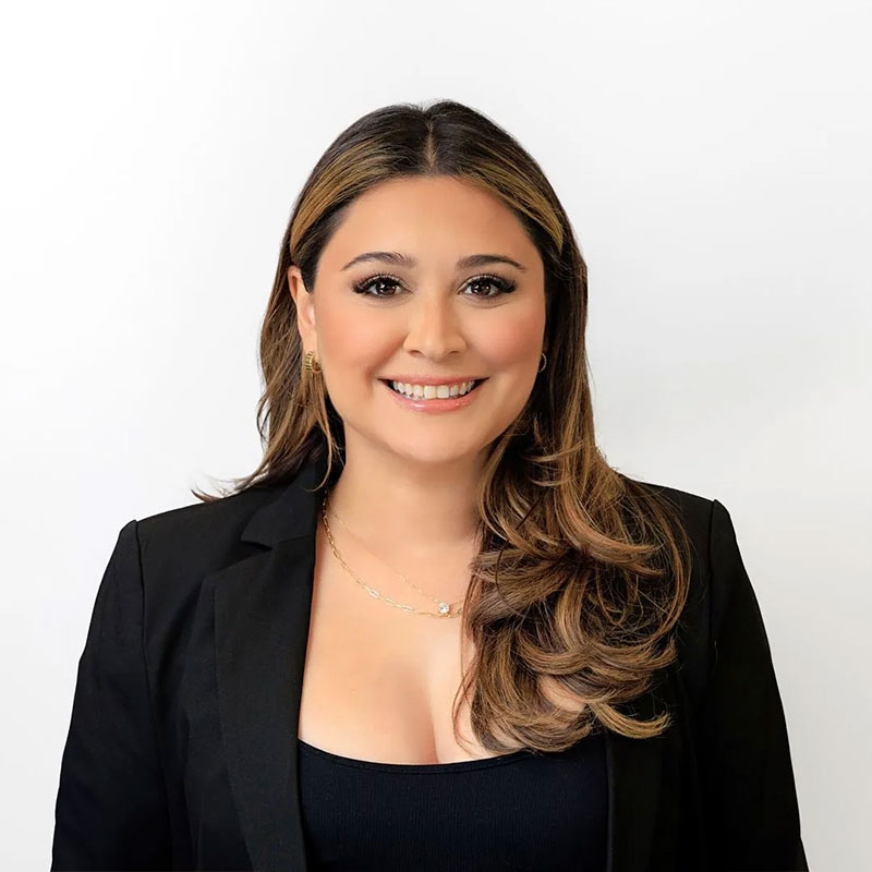 Smiling woman with long wavy hair wearing a black blazer and necklace on white background.