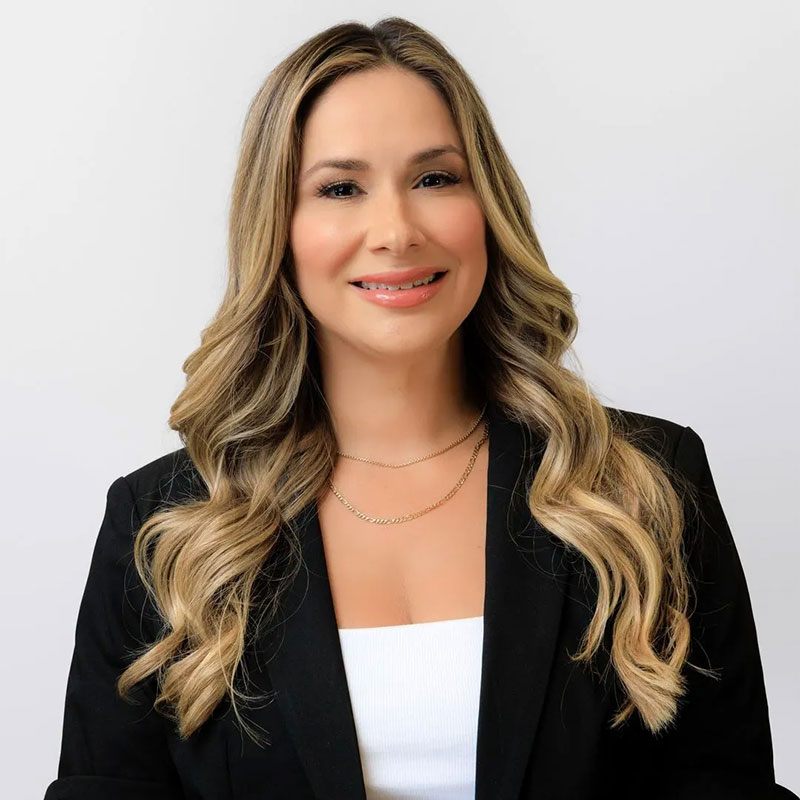 Smiling woman with long wavy blonde hair wearing a black blazer and white top against plain background.