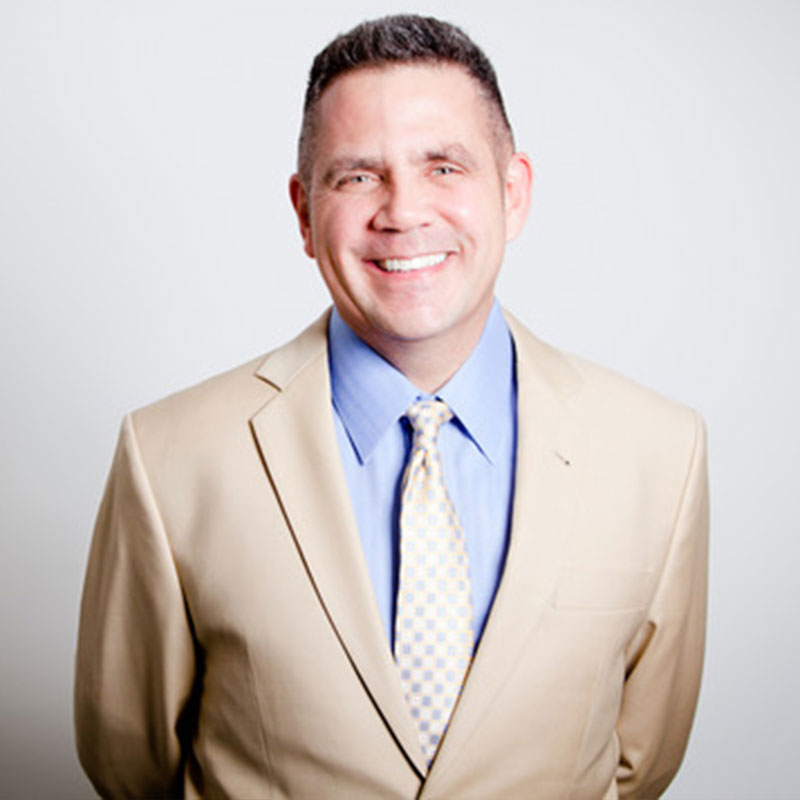 Man smiling wearing beige suit jacket, blue shirt, and patterned tie against a light background.