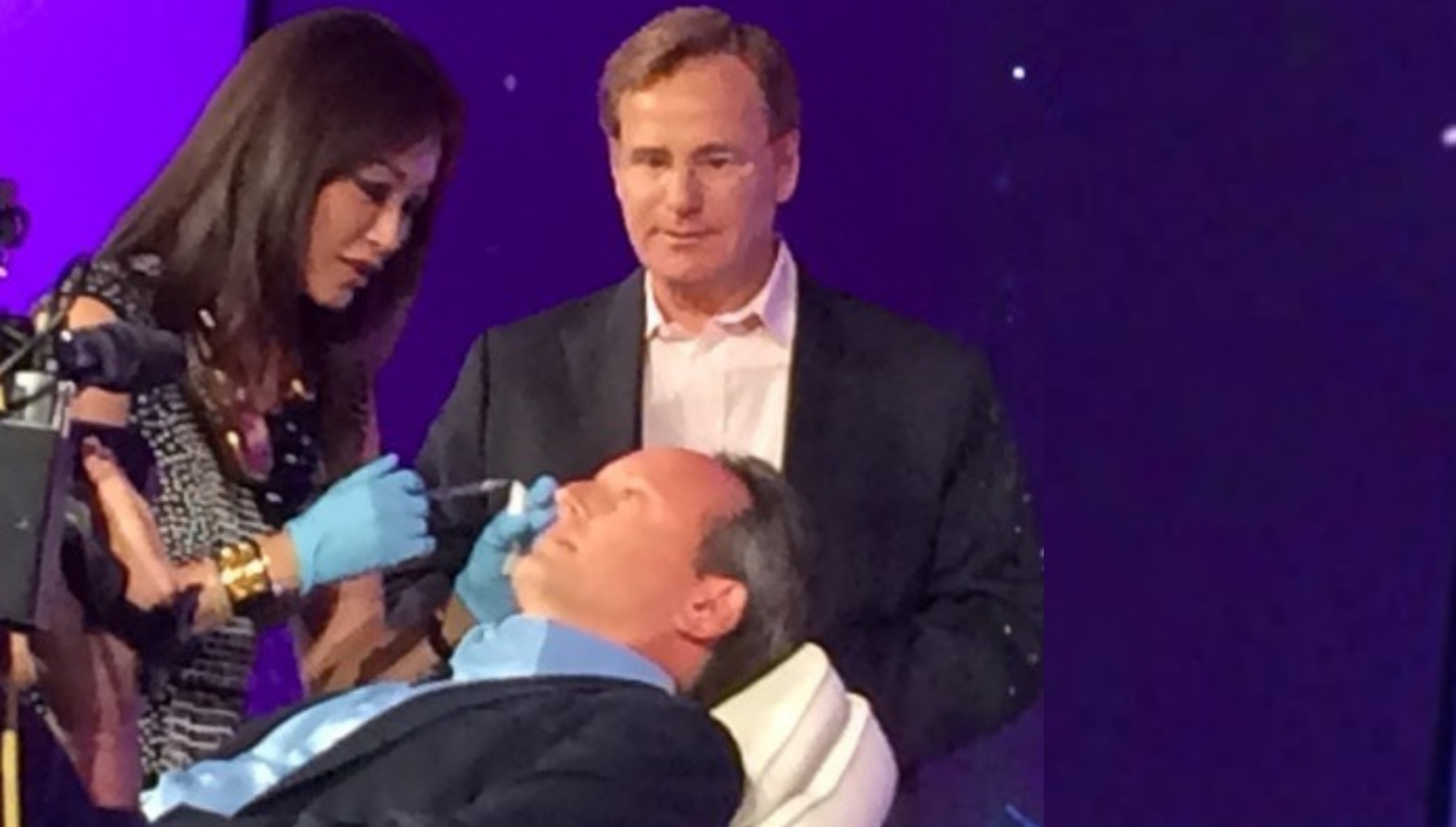 Woman wearing gloves performs a facial treatment on a seated man while another man watches.