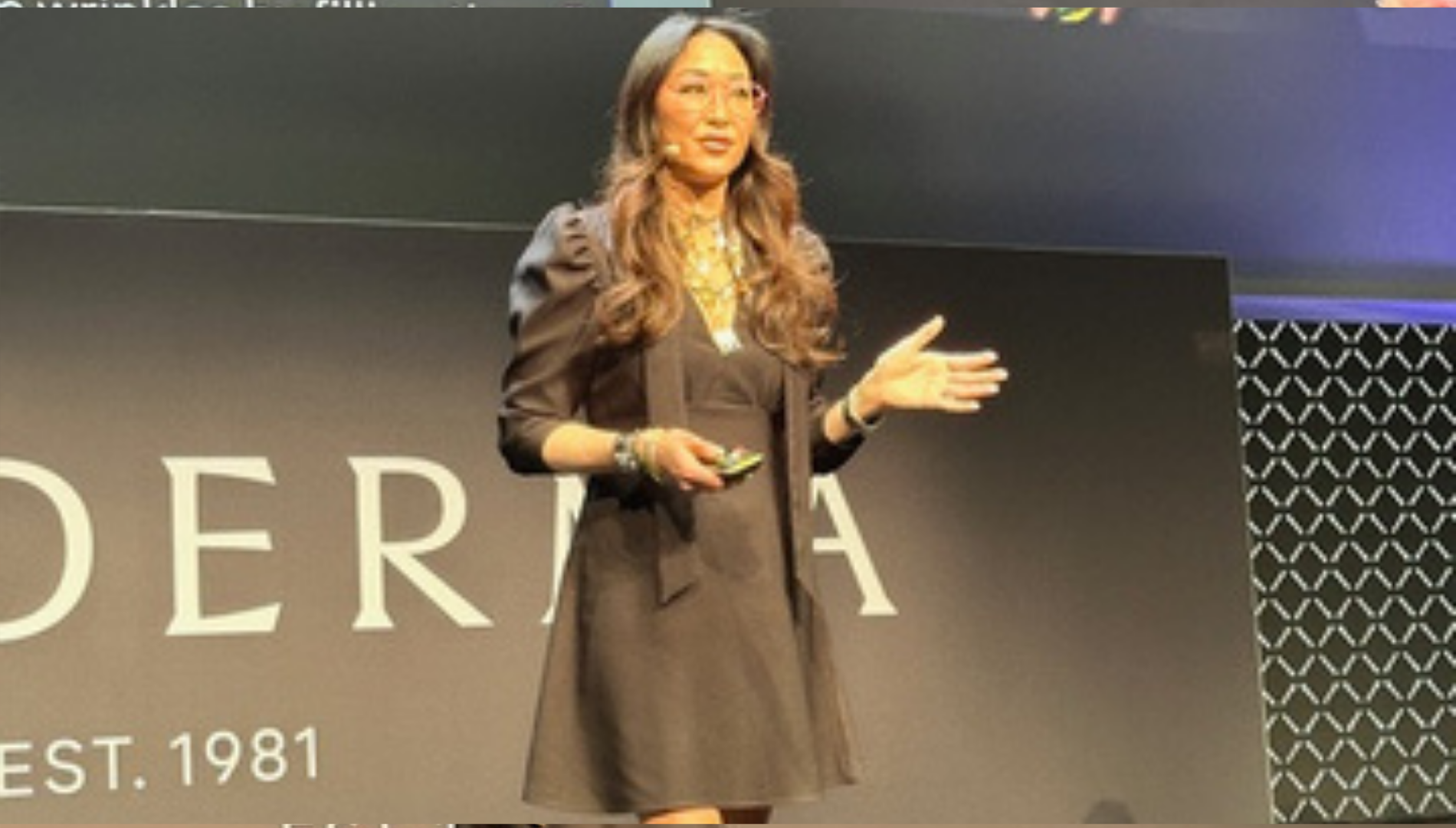 Woman with long hair and glasses speaking onstage wearing a black dress and holding a remote.