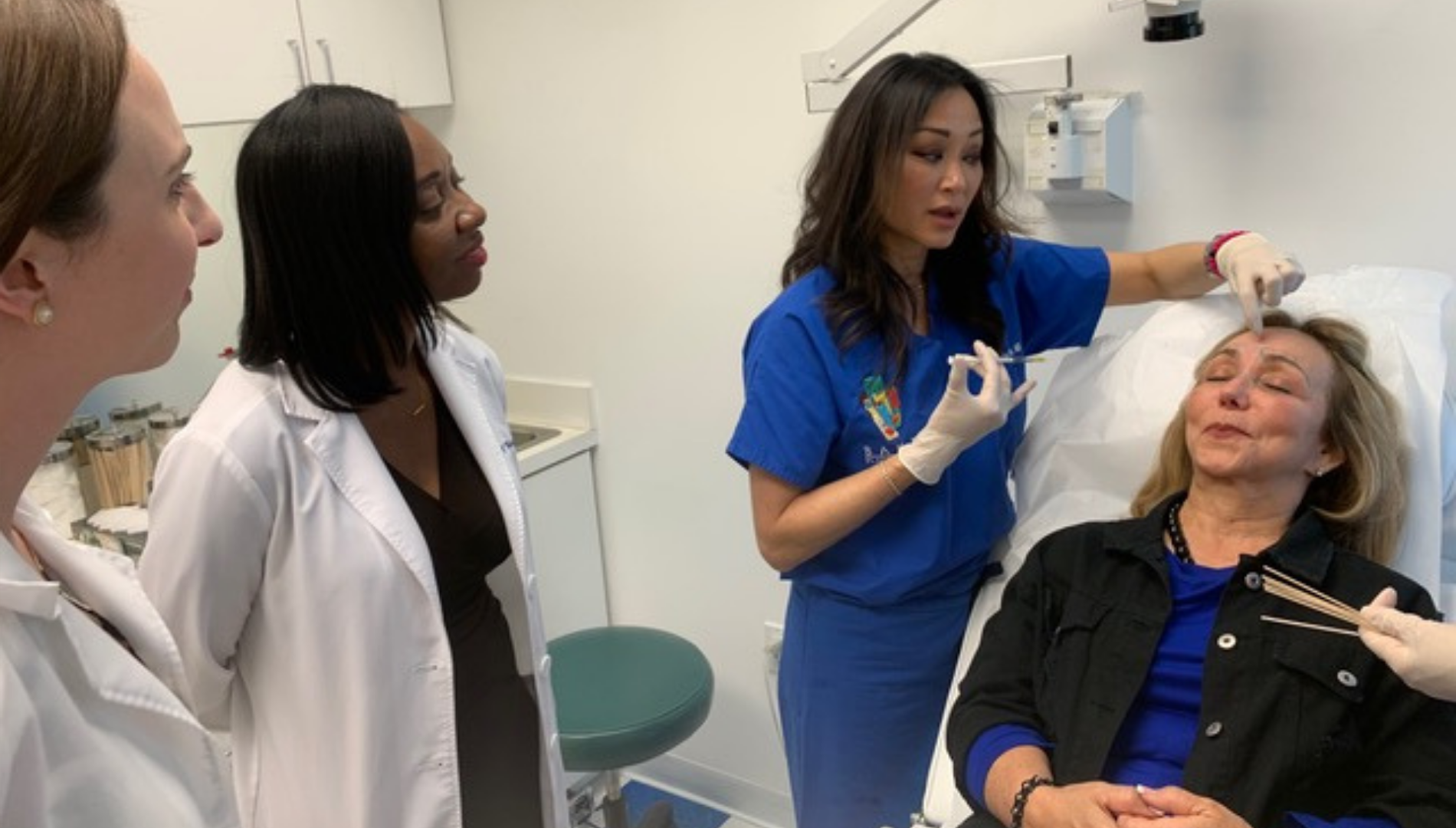 Healthcare professional in blue scrubs explains injection on woman’s forehead to two observers in white lab coats.