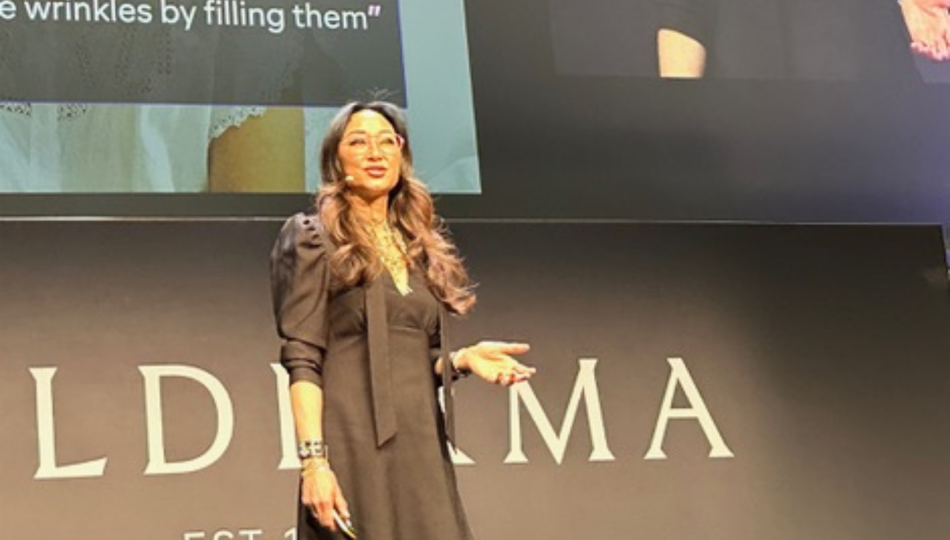 Woman with long hair and glasses speaking on stage in a black dress with puffed sleeves.