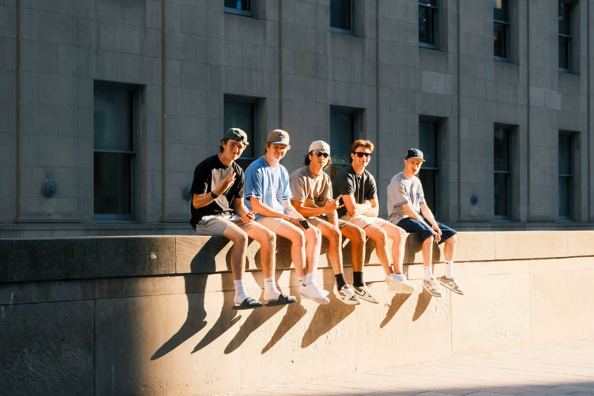 Five young men in casual summer attire sit on a stone ledge, smiling and relaxed. The warm sunlight casts long shadows on the wall behind them.
