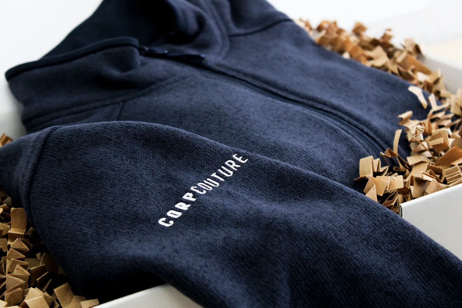 Dark navy zip-up fleece jacket with the word 'CORPCOUTURE' embroidered on the sleeve, resting in a box with brown packing paper.