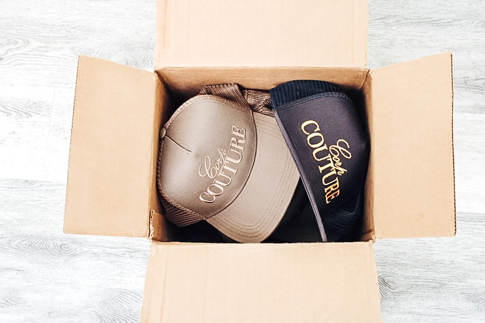 Two Corporate Couture baseball caps, one brown and one navy, inside an open cardboard box on a light wooden floor.