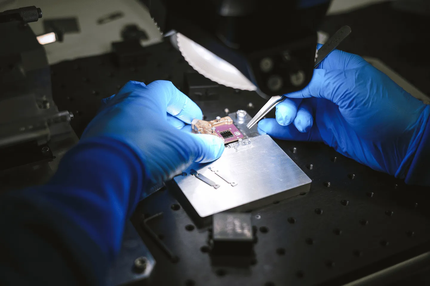 Person wearing blue gloves assembling or inspecting a small electronic chip on a metal platform with tweezers under a microscope light.