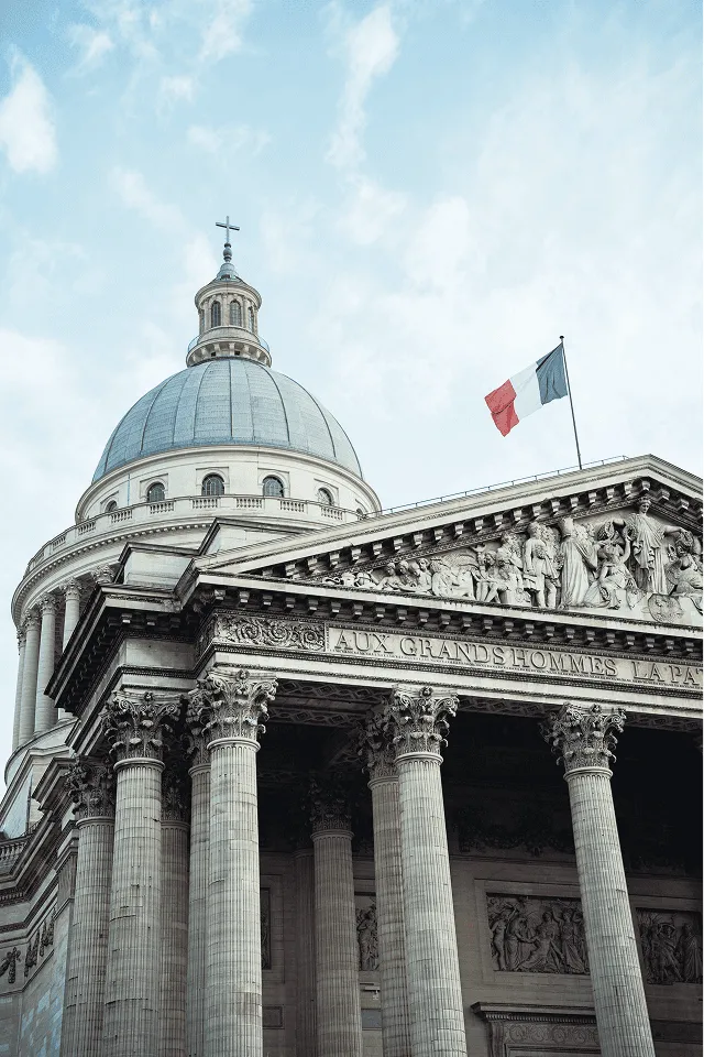 Low-angle view of the Pantheon in Paris with a French flag flying above its Corinthian columns and dome under a cloudy sky.