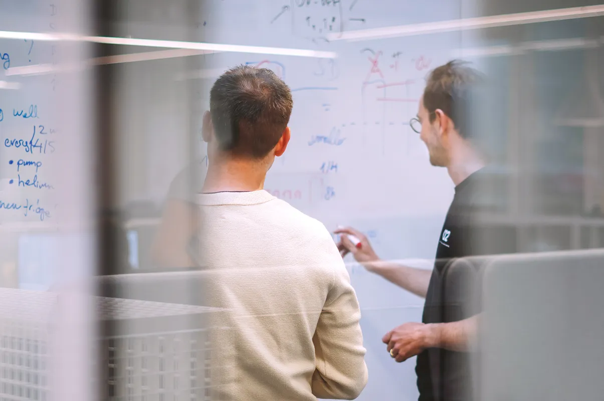 Two men discussing and writing on a whiteboard filled with notes and diagrams in an office.