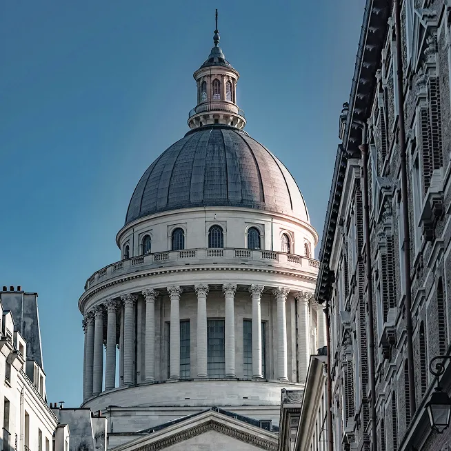 Close-up view of a large classical dome with columns illuminated by sunlight against a clear blue sky, flanked by older buildings on both sides.