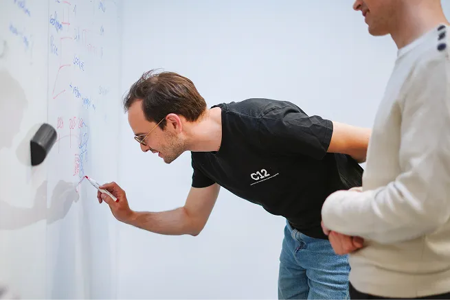 Two men collaborating at a whiteboard, one writing with a marker while the other observes.