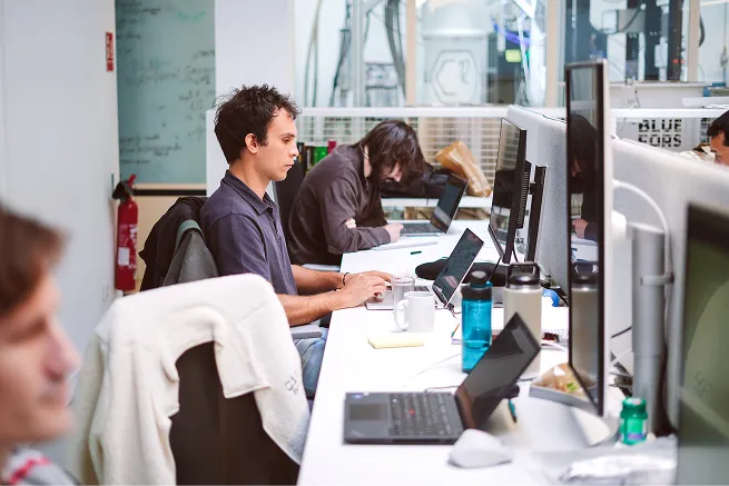Young professionals working on laptops at a shared office desk in a modern workspace.