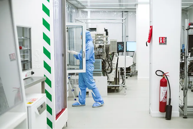Person wearing blue cleanroom suit working in a sterile laboratory with scientific equipment and fire extinguisher on the wall.