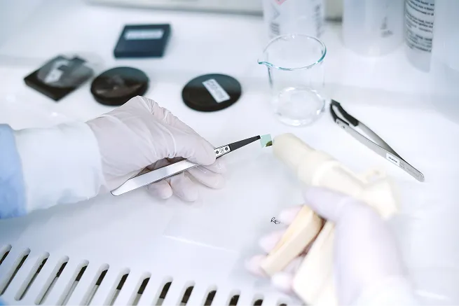 Gloved hands using tweezers to hold a small green test strip in a laboratory setting with scientific tools and containers on the table.