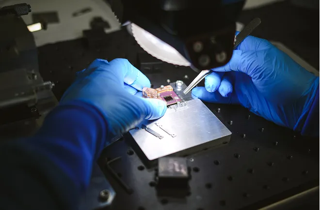 Person wearing blue gloves handling a small circuit board with tweezers under a focused light on a workbench.