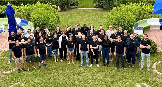 Group of 30 adults standing outdoors on grass, smiling and facing the camera, wearing mostly black shirts, with bushes and curved benches in the background.