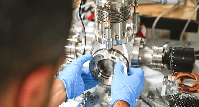 Person wearing blue gloves handling a metal component of a laboratory instrument.