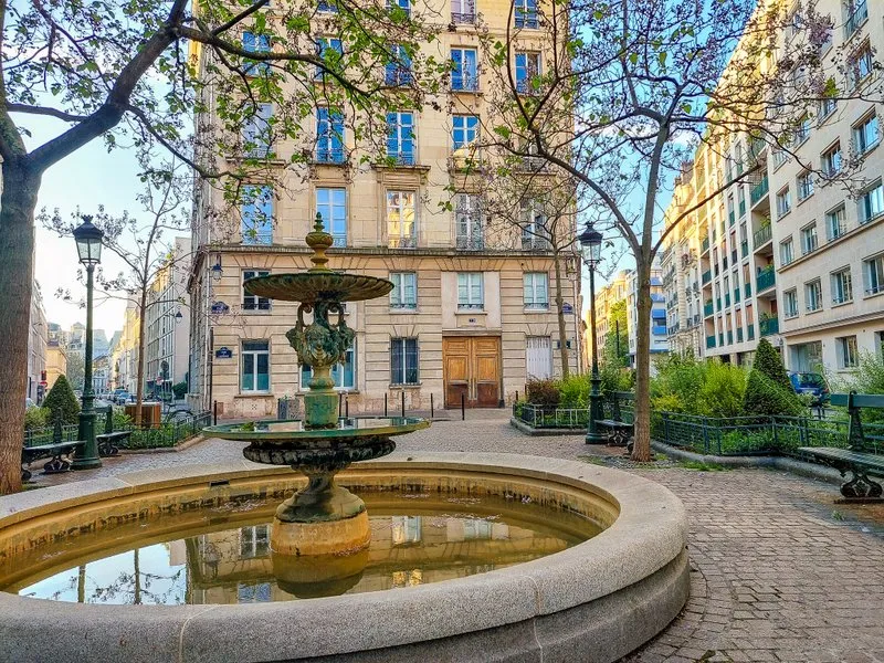 Stone fountain with water in the foreground of a cobblestone street and beige building with tall windows and wooden door.