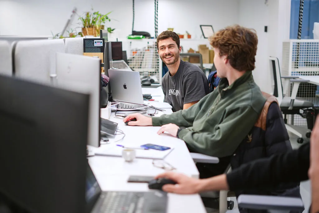 Man in striped sweater working on a laptop at a shared office desk with computer monitors.