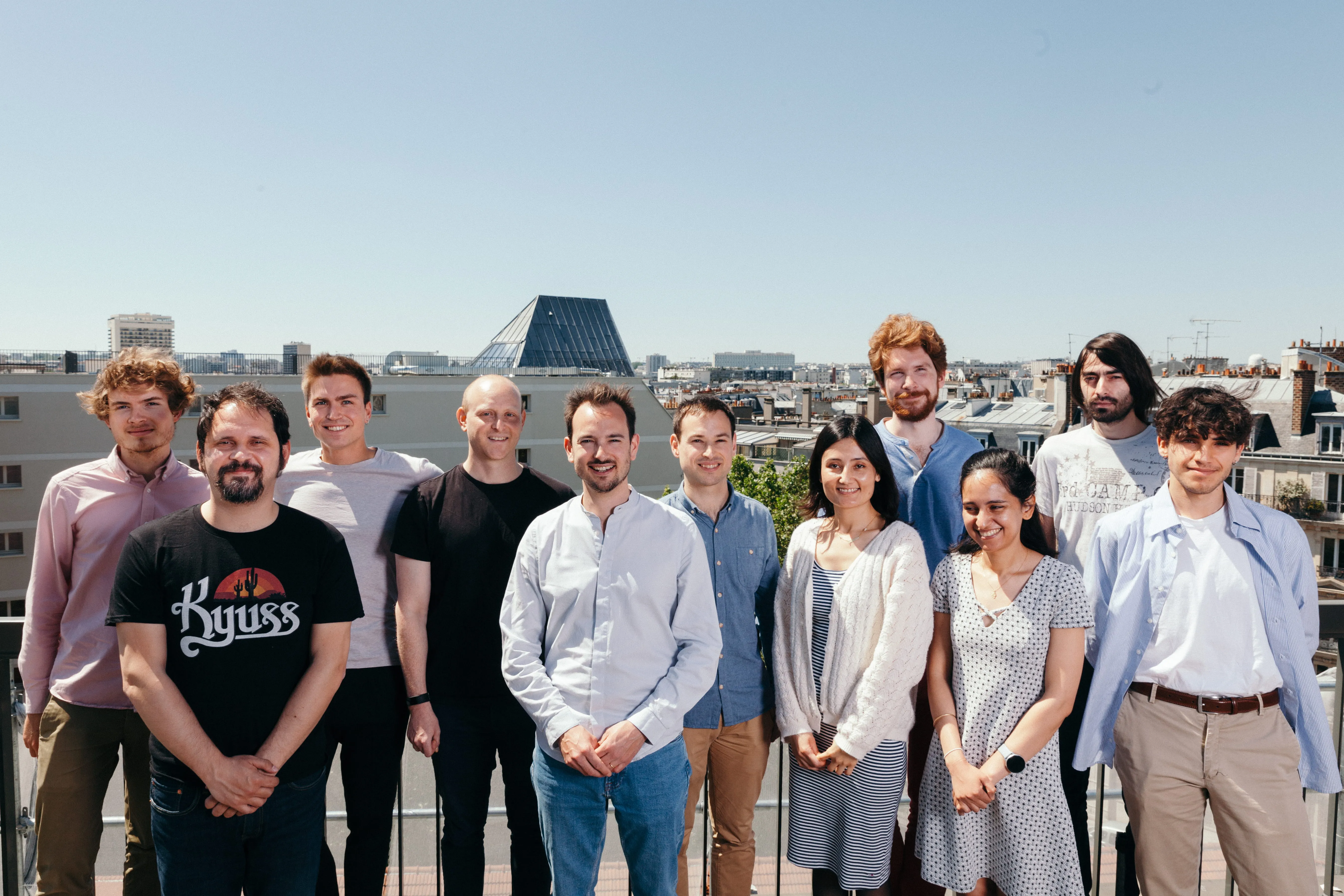 Group of eleven men and women standing outdoors with a cityscape in the background under a clear sky.