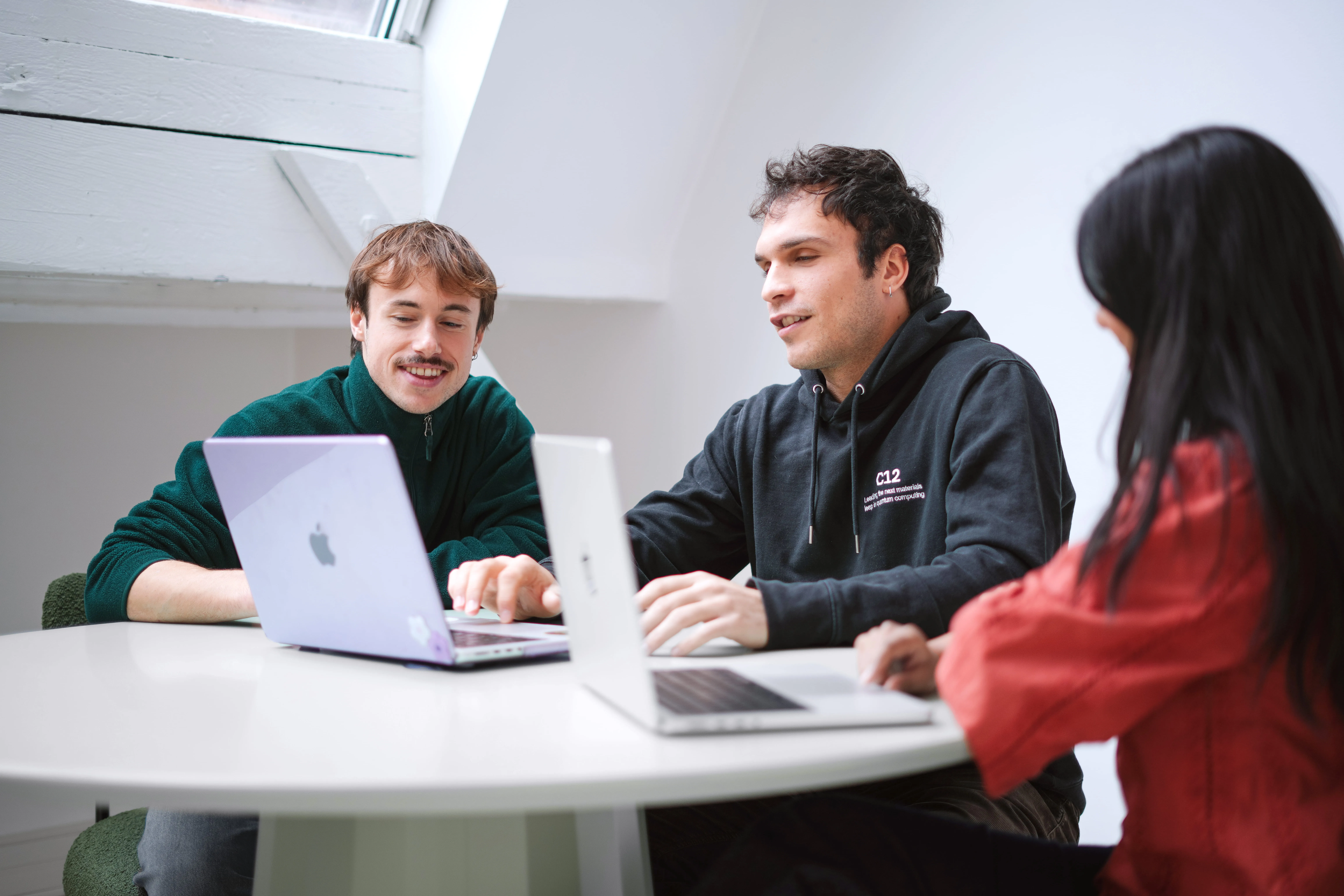 Three young adults sitting around a white round table, working on laptops and engaging in a discussion in a bright room.