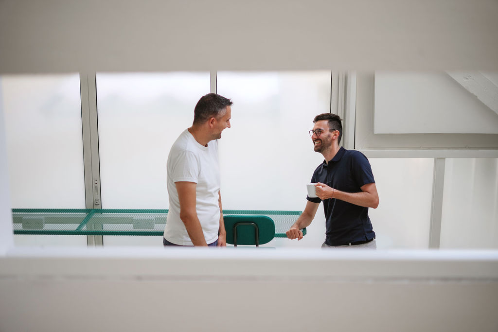 Modern office kitchen and meeting area with people sitting at a long table and one person walking in the background.