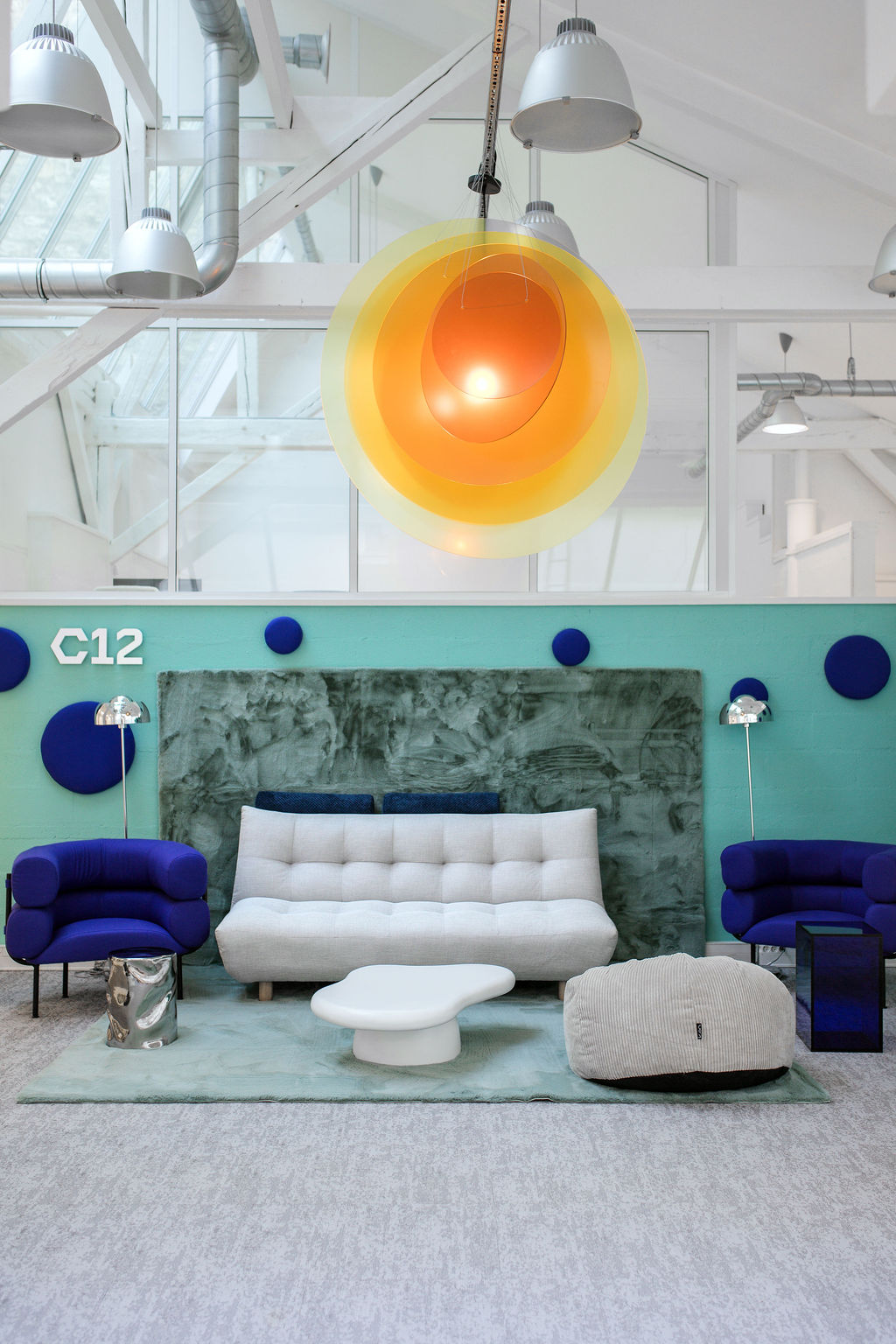 Modern office lounge with a white sofa, blue armchair, large yellow-orange hanging lamp, and textured turquoise wall labeled C12.