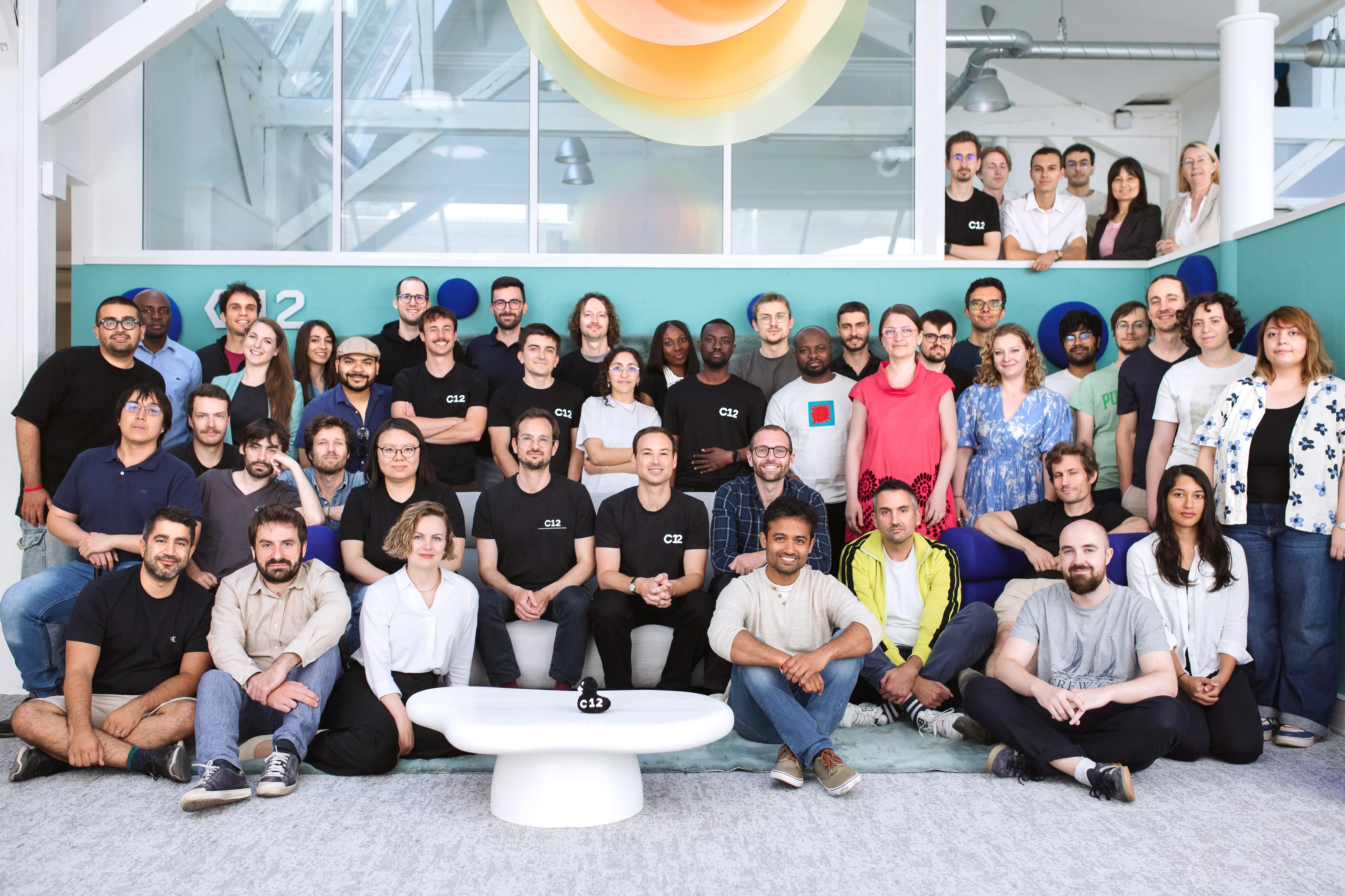 Large diverse group of people posing indoors in front of a teal wall with a white table in front.