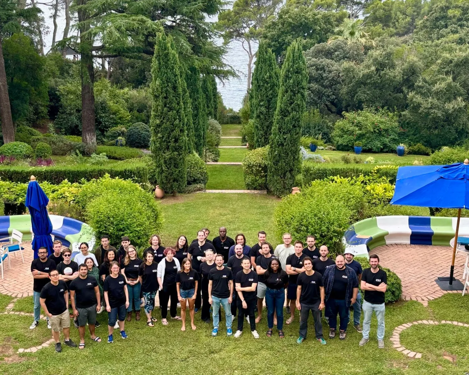 Group of 30 adults standing outdoors on grass, smiling and facing the camera, wearing mostly black shirts, with bushes and curved benches in the background.