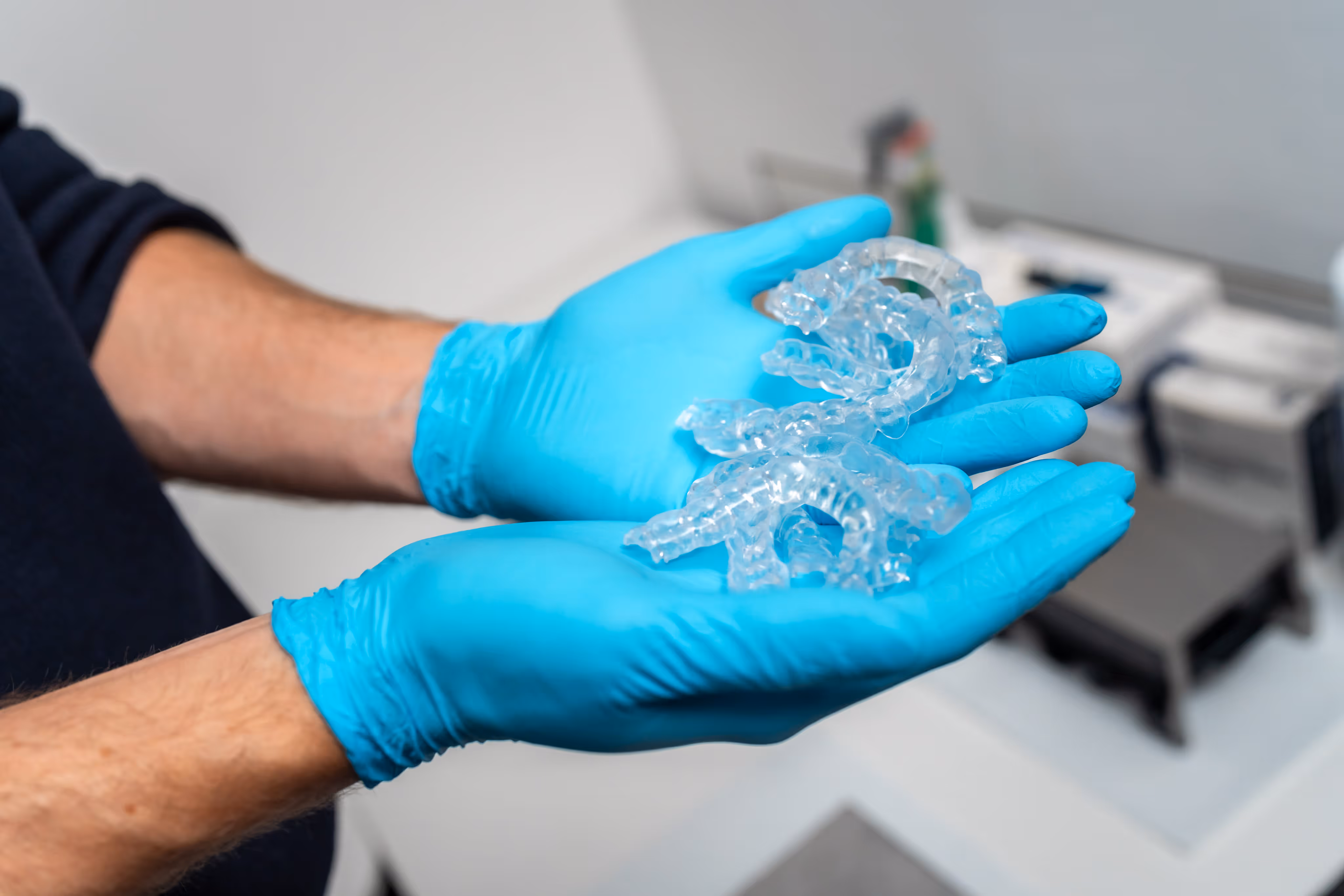 Hands in blue gloves holding multiple clear dental aligners in a clinical setting.