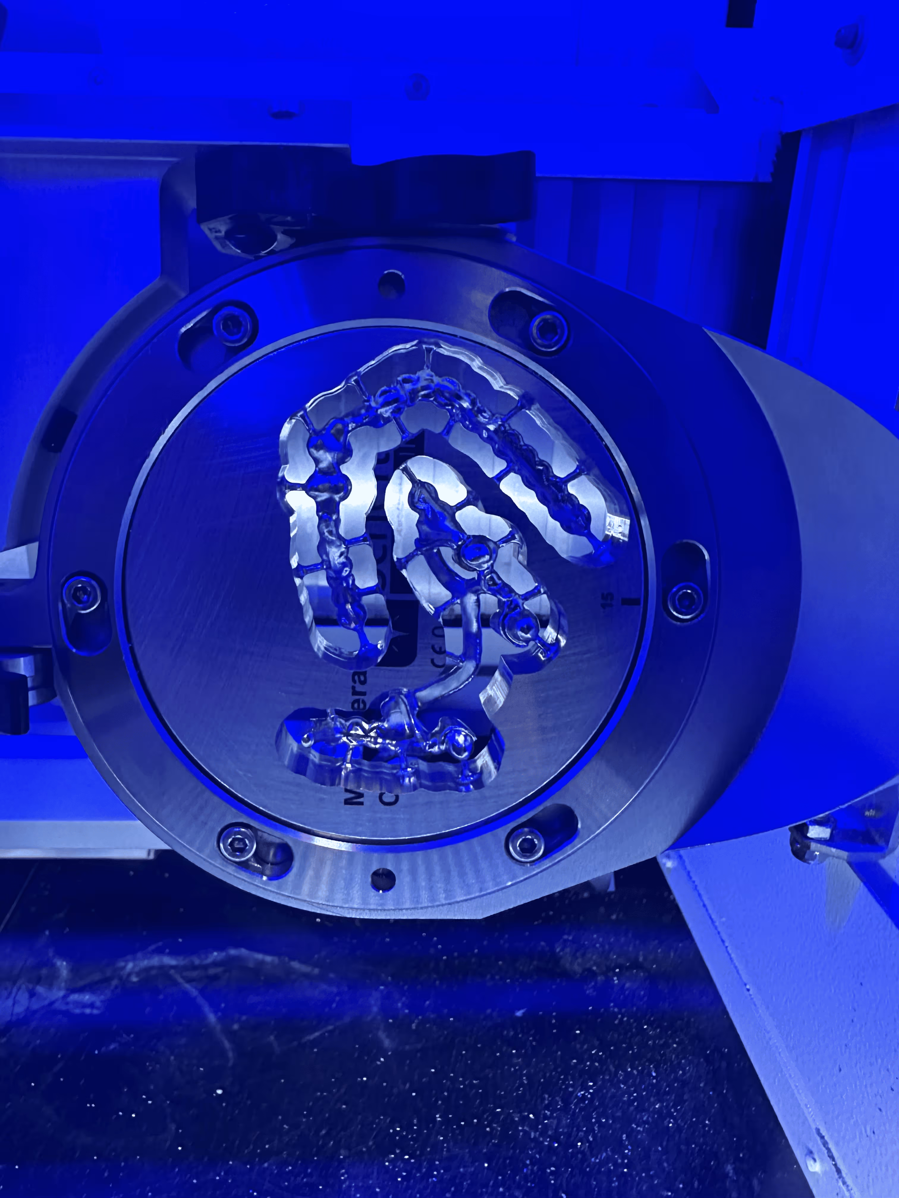 Clear dental aligner attached to a metal circular component in a machine illuminated by blue light.