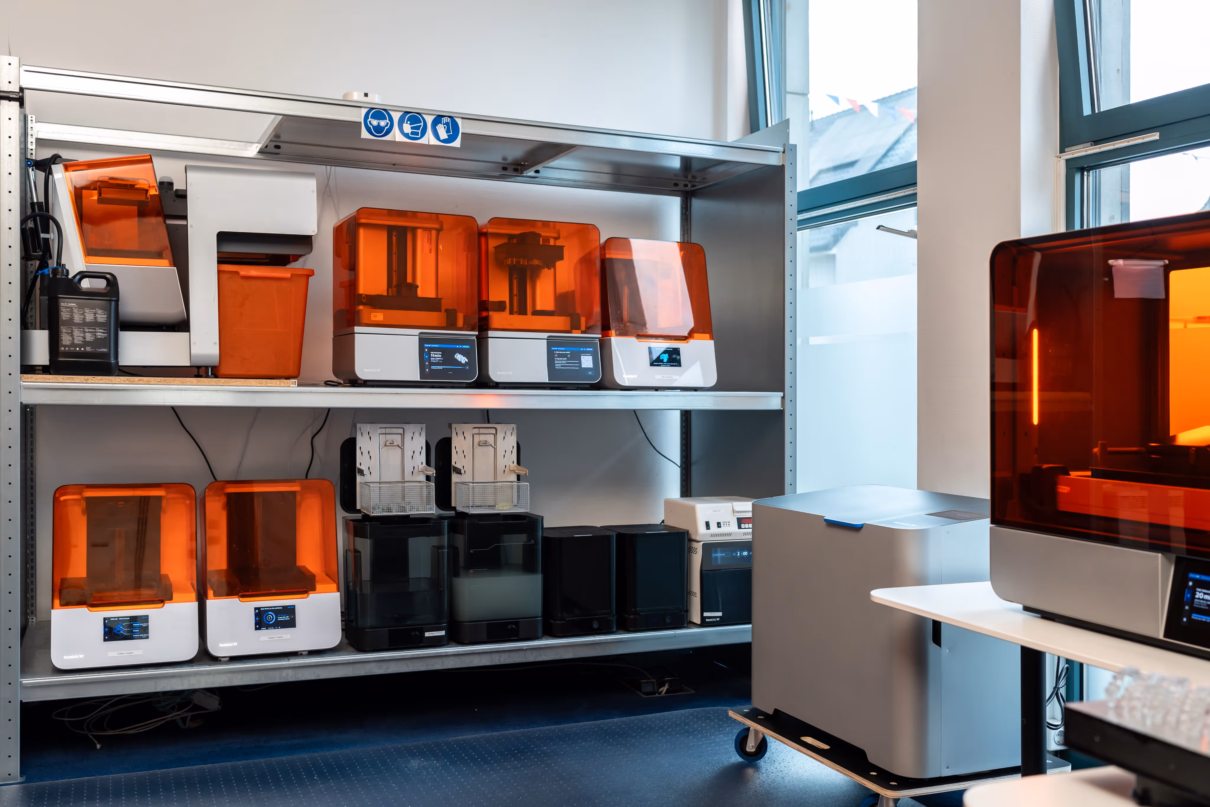 Shelves with multiple orange-covered resin 3D printers and lab equipment in a bright room with large windows.