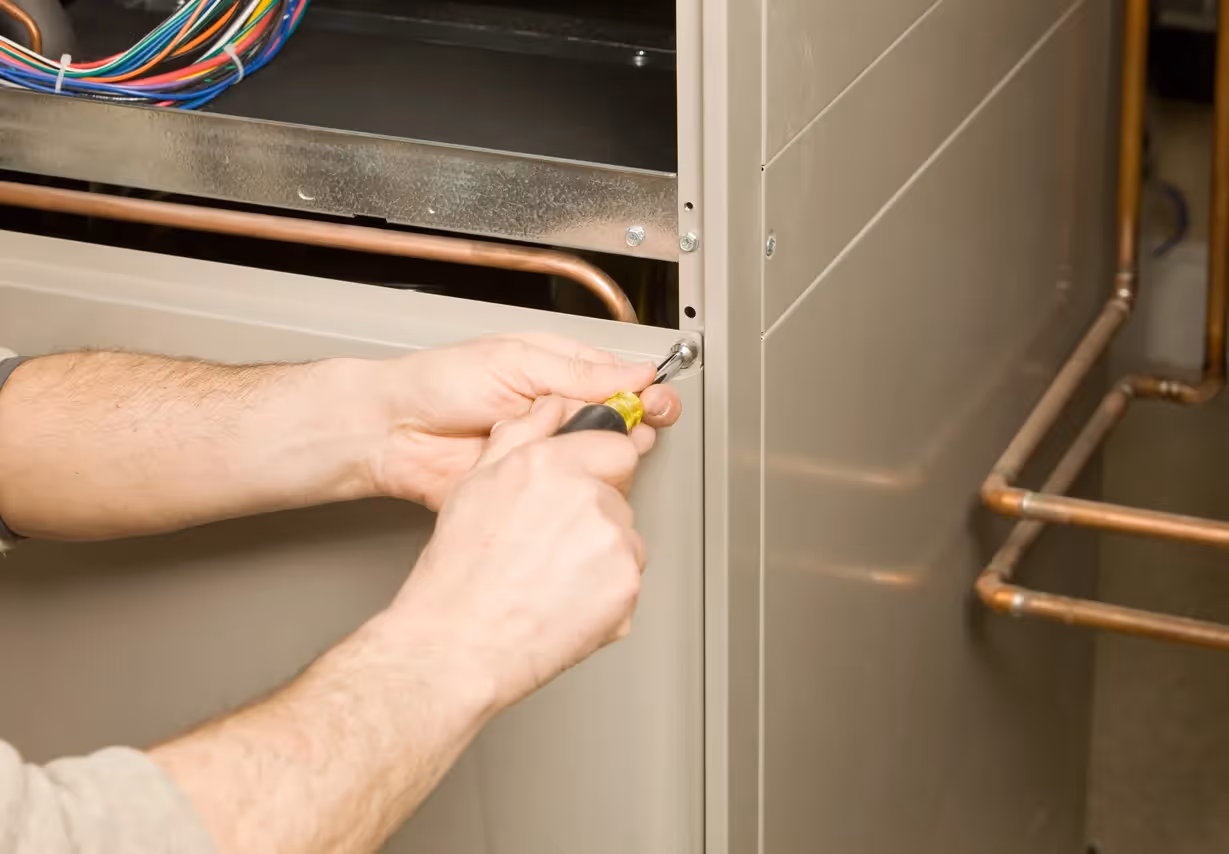 A close-up shot of a person using a screwdriver to remove a screw from the metal casing of a furnace, part of a home repair or maintenance task.