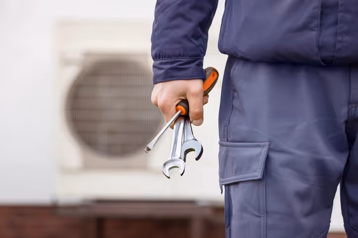 A technician in blue workwear holds a screwdriver and wrenches in front of an air conditioner