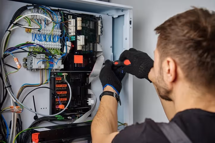 Technician wearing black gloves uses a screwdriver to adjust wiring inside an electrical control panel with multicolored cables and components.