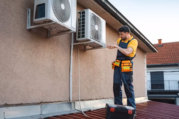 Technician in yellow shirt and blue overalls stands on sloped metal roof servicing wall-mounted AC units near toolbox.