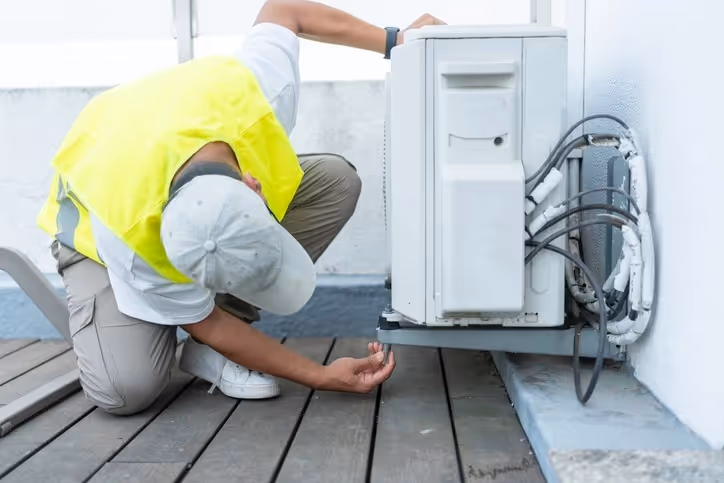Technician in yellow safety vest inspects outdoor AC unit on rooftop, reaching beneath and touching top.