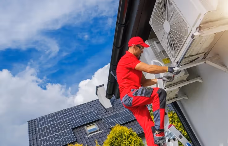 Technician in red uniform and safety glasses stands on ladder servicing wall-mounted AC unit, solar panels on dark tiled roof.