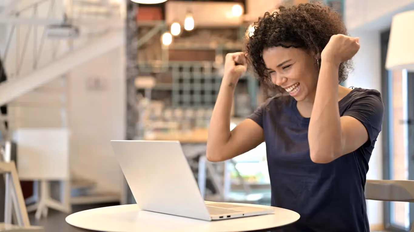 A happy woman with curly hair cheering in front of her laptop in a bright office