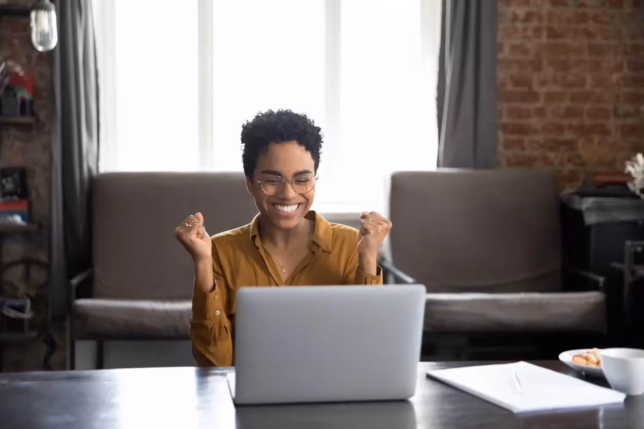 A woman wearing glasses celebrates with raised fists while looking at her laptop in a living room
