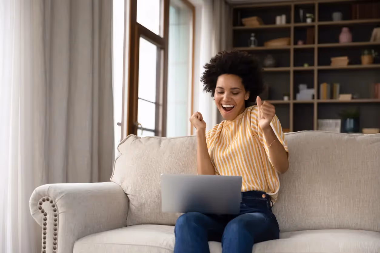A woman sitting on a sofa laughs and cheers while looking at her laptop screen
