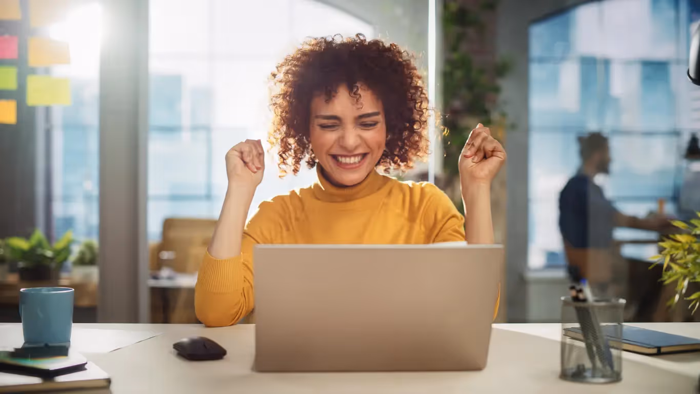 A smiling woman in a yellow sweater cheers excitedly at her laptop in a sunlit office