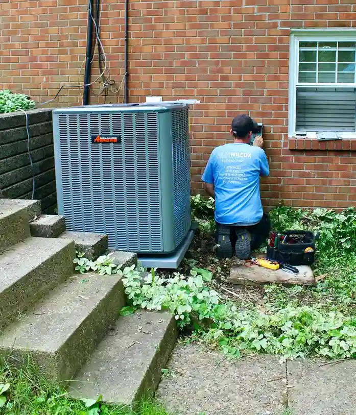 An air conditioner technician kneeling to work on a large outdoor AC unit next to a brick building.