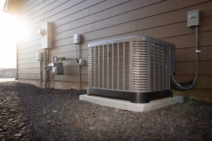 A residential air conditioning unit rests on a gravel bed next to a house, with utility meters and pipes visible.